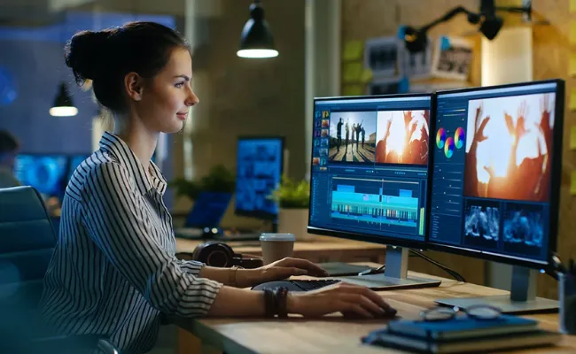 Female video editor working on dual monitors to color grade footage, showcasing professional video color grading and video post-production services by a top video editing agency.