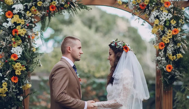 Bride and groom holding hands under a flower-decorated arch during an outdoor wedding ceremony, showcasing natural light ideal for wedding videography