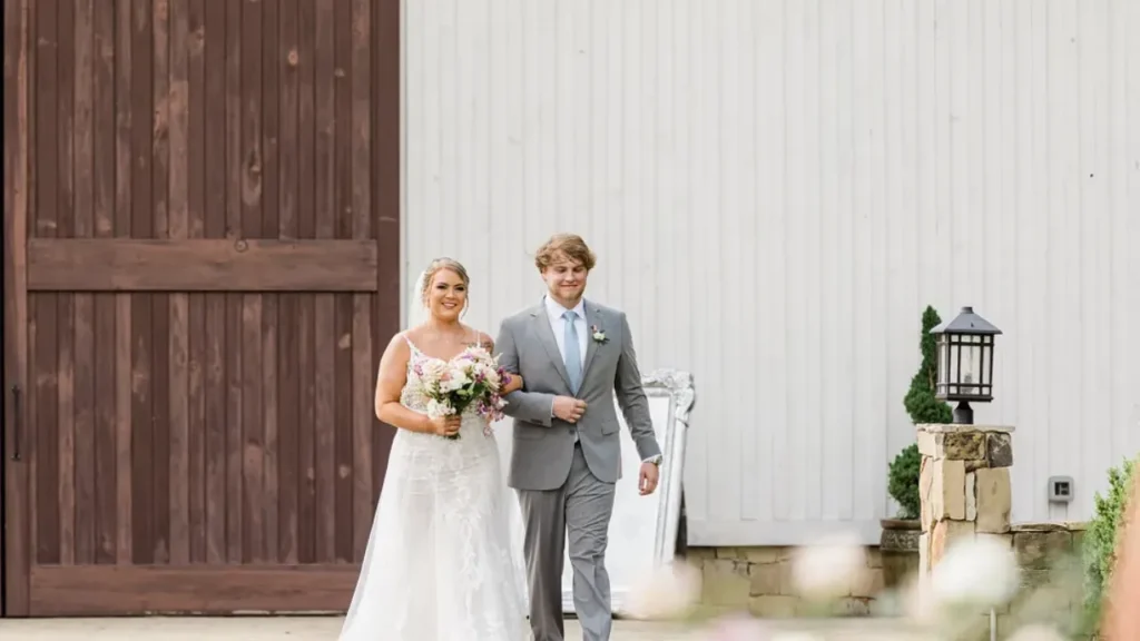 Bride walking arm-in-arm with a groomsman in front of rustic barn doors, capturing a key emotional moment perfect for storytelling in a cinematic wedding highlight video