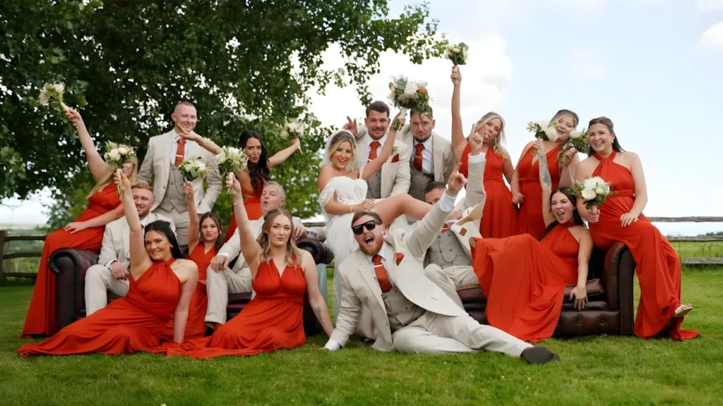 Bride and groom posing with joyful bridesmaids and groomsmen in coordinated red and beige outfits, capturing vibrant candid moments perfect for wedding highlight video and storytelling highlights for wedding